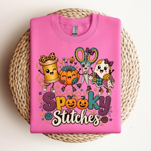 Peut inclure: Un sweat-shirt à col rond rose vif avec un motif coloré représentant une bobine de fil, un coussin à épingles citrouille, des ciseaux et un fantôme, tous avec des visages. Le texte indique Spooky Stitches avec deux petites citrouilles.