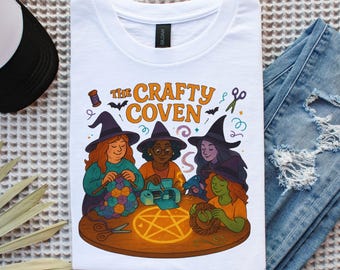 May include: White t-shirt with the words "The Crafty Coven" and illustrations of three witches crafting. The witches have orange, green, and purple hair, wear black witch hats, and are engaged in different crafts. Scissors and a pentagram are also shown.