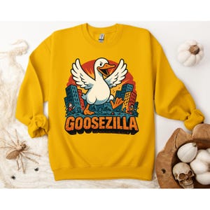 Puede incluir: Sudadera amarilla mostaza con un gráfico de dibujos animados de un ganso llamado "GOOSEZILLA" pisoteando una ciudad. El ganso es blanco con pico y patas naranjas. Los edificios son azules y naranjas.