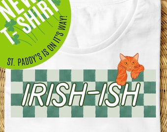 May include: White t-shirt with a green and white checkered pattern and the text "IRISH-ISH" in white. A cartoon orange cat is peeking over the top of the checkered pattern.
