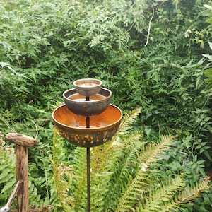 May include: A three-tiered metal bird feeder with rusty brown and grey bowls. The feeder is mounted on a tall black metal pole and is surrounded by green ferns and foliage.