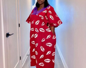 May include: A red midi dress with white lip print. The dress features a purple neckline and short sleeves. The dress has pockets and is worn by a person.