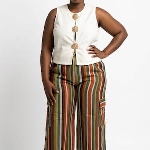 May include: A sleeveless, off-white vest with three decorative gold buttons is paired with wide-leg, striped pants. The pants feature vertical stripes in shades of brown, green, and orange. The model is wearing white sandals.