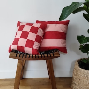 Peut inclure: Deux coussins décoratifs sur un tabouret en bois. Un coussin est à carreaux rouge et blanc, l'autre est à rayures rose et rouge.