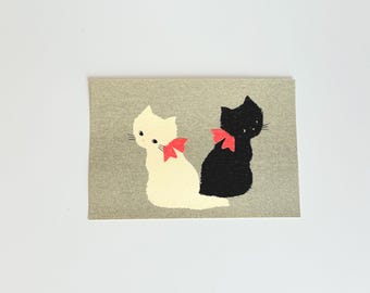 May include: A rectangular card with a design of two cats. One is white, the other black, each with a red bow. They sit together on a textured grey background. The illustration has a simple, charming aesthetic, reminiscent of a children's book.