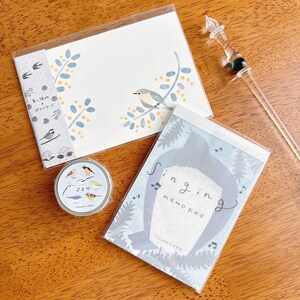 May include: A collection of stationery items featuring bird illustrations. Includes a notepad with the text "Singing memo pad", a postcard, and a roll of decorative tape. The items are in a clear plastic packaging.