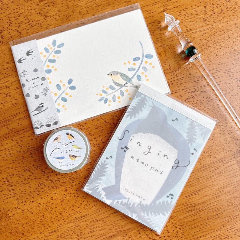 May include: A collection of stationery items featuring bird illustrations. Includes a notepad with the text "Singing memo pad", a postcard, and a roll of decorative tape. The items are in a clear plastic packaging.
