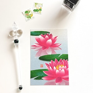 May include: A postcard featuring a pink lotus flower with green lily pads, reflecting in the water. A clear glass pen and two stamps are also visible. The stamps have a floral design and the number 110.