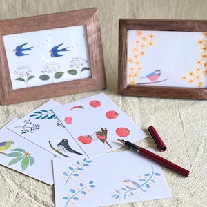 May include: Two framed art prints with nature-themed illustrations, including birds and floral designs. Several art cards with bird and botanical illustrations are arranged in front of the frames. A burgundy fountain pen is also visible.