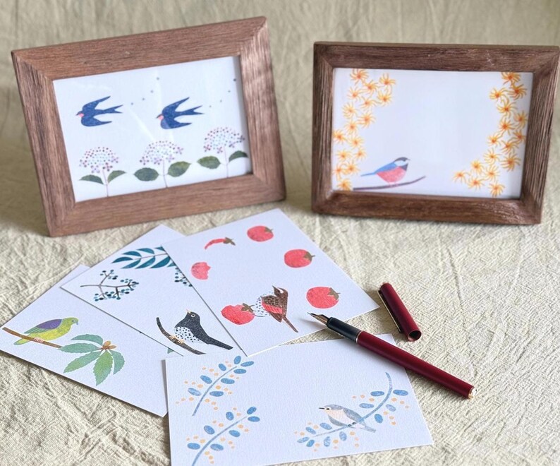 May include: Two framed art prints with nature-themed illustrations, including birds and floral designs. Several art cards with bird and botanical illustrations are arranged in front of the frames. A burgundy fountain pen is also visible.