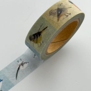 May include: Decorative washi tape featuring a repeating pattern of various birds. The tape has a light blue and green background with illustrations of birds in different poses. The tape is rolled up, showing the end of the roll and a portion of the unrolled tape.