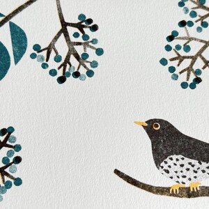 May include: A detailed illustration of a black bird with white speckled markings perched on a branch. The artwork features teal-colored berries and leaves against a white background, creating a nature-inspired design.