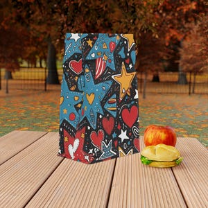 Op de afbeelding: Een kleurrijke herbruikbare lunchtas met een zwarte achtergrond en een patroon van blauwe sterren, rode harten en gele sterren. De tas is gemaakt van duurzaam materiaal en is perfect voor het dragen van snacks en drankjes.