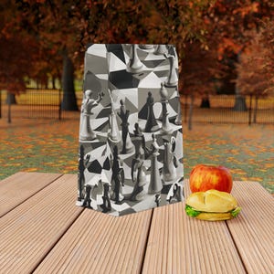 May include: A brown paper bag with a black and white chessboard pattern featuring chess pieces. The bag is sitting on a wooden surface with a red apple and a sandwich.