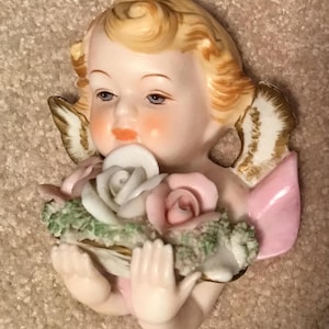 May include: A ceramic wall plaque featuring a cherub with blonde hair and gold-trimmed wings. The cherub holds a bouquet of sculpted white and pink roses accented with green foliage. The figure has a pale complexion, rosy cheeks, and orange lips.