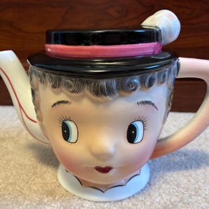 May include: A ceramic teapot shaped like a face with a black hat lid featuring a pink band. The teapot has a white spout, a pink handle, and painted features including blue eyes, dark hair, and red lips. The lid includes a small white accent.