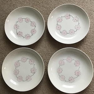 May include: Four white ceramic plates with a floral design. Each plate features a ring of pink and gray stylized flowers around the center. The plates are round and have a slightly raised edge.