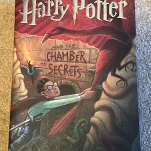 May include: A hardcover book titled Harry Potter and the Chamber of Secrets by J.K. Rowling. The cover features a young person with glasses reaching toward a large red bird perched on a stone structure, with a brick wall background and text.