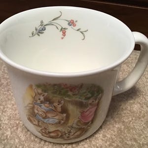 May include: White ceramic mug with a large handle. The mug features a colorful illustration of rabbits in a garden scene. Delicate floral sprigs with blue and red accents adorn the rim. A charming and whimsical design for a tea or coffee cup.