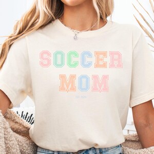 May include: A white t-shirt with the text "SOCCER MOM" in pastel colors. The letters are outlined in a slightly darker shade of the same color. The text "EST. 2022" is printed in a smaller font below the main text.