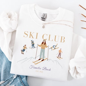 Op de afbeelding: Wit sweatshirt met een afbeelding van skiërs en de tekst "SKI CLUB Familie Bach EST. 2023"