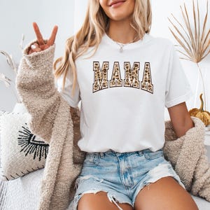 May include: White t-shirt with the word "MAMA" printed in brown and tan leopard print letters.