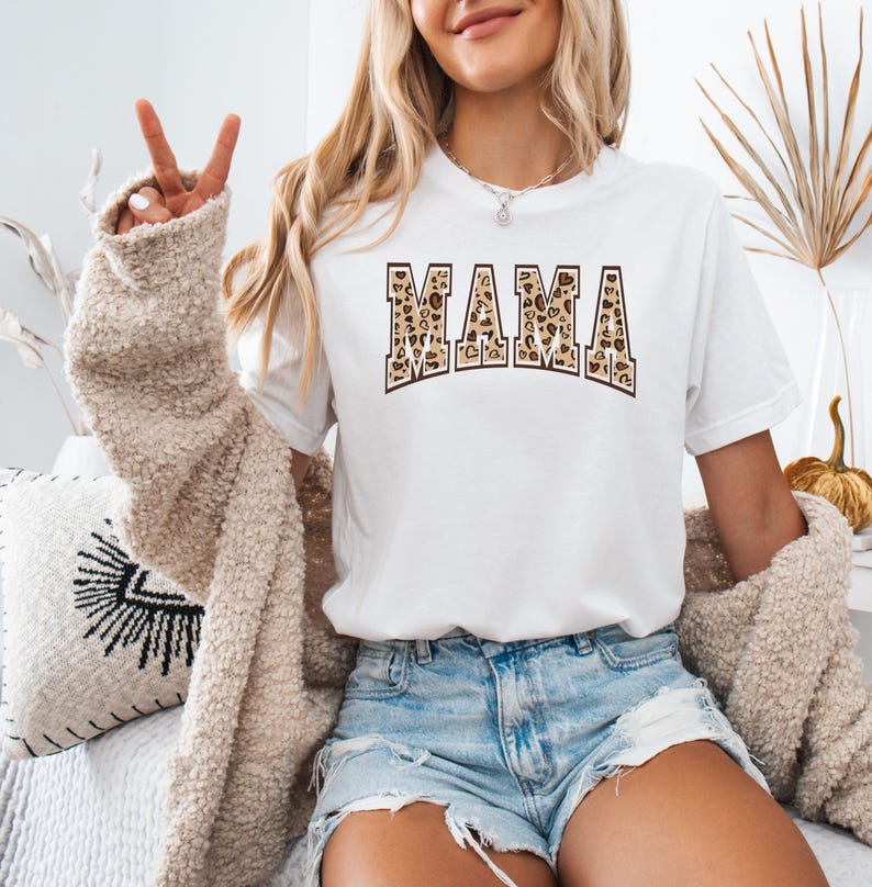 May include: White t-shirt with the word "MAMA" printed in brown and tan leopard print letters.