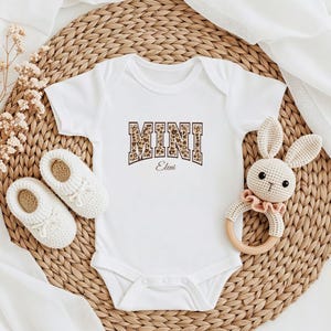 May include: White baby onesie with a brown and white leopard print "MINI" graphic with the name "Eleni" underneath. The onesie is on a woven straw mat with a crocheted bunny toy and a pair of white baby shoes.