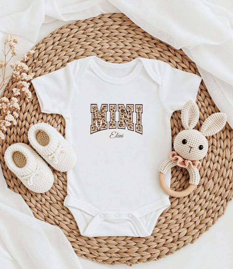 May include: White baby onesie with a brown and white leopard print "MINI" graphic with the name "Eleni" underneath. The onesie is on a woven straw mat with a crocheted bunny toy and a pair of white baby shoes.
