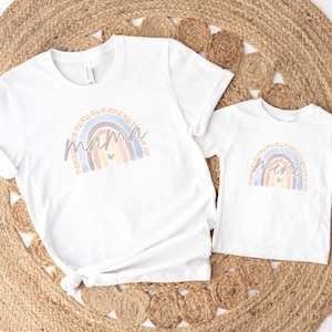 May include: Two white t-shirts with a rainbow design and the words "mama" and "mini" on them. The shirts are laid out on a woven rug.