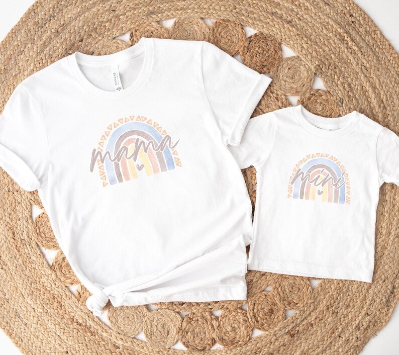 May include: Two white t-shirts with a rainbow design and the words "mama" and "mini" on them. The shirts are laid out on a woven rug.