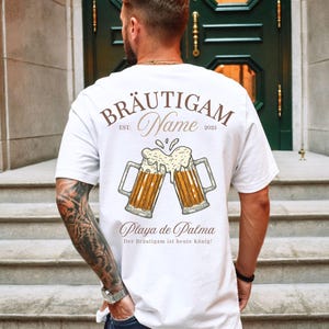 Puede incluir: Camiseta blanca con un dise&ntilde;o de tem&aacute;tica cervecera. El dise&ntilde;o incluye dos jarras de cerveza chocando, con las palabras "BR&Auml;UTIGAM Name EST. 2025" y "Playa de Palma" impresas en la espalda. Tambi&eacute;n se incluye el texto "Der Br&auml;utigam ist heute K&ouml;nig!"