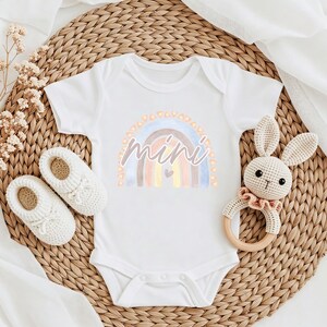 May include: White baby onesie featuring a watercolour rainbow design and the word "mini" in a pink script font. The rainbow has a small heart in the centre.