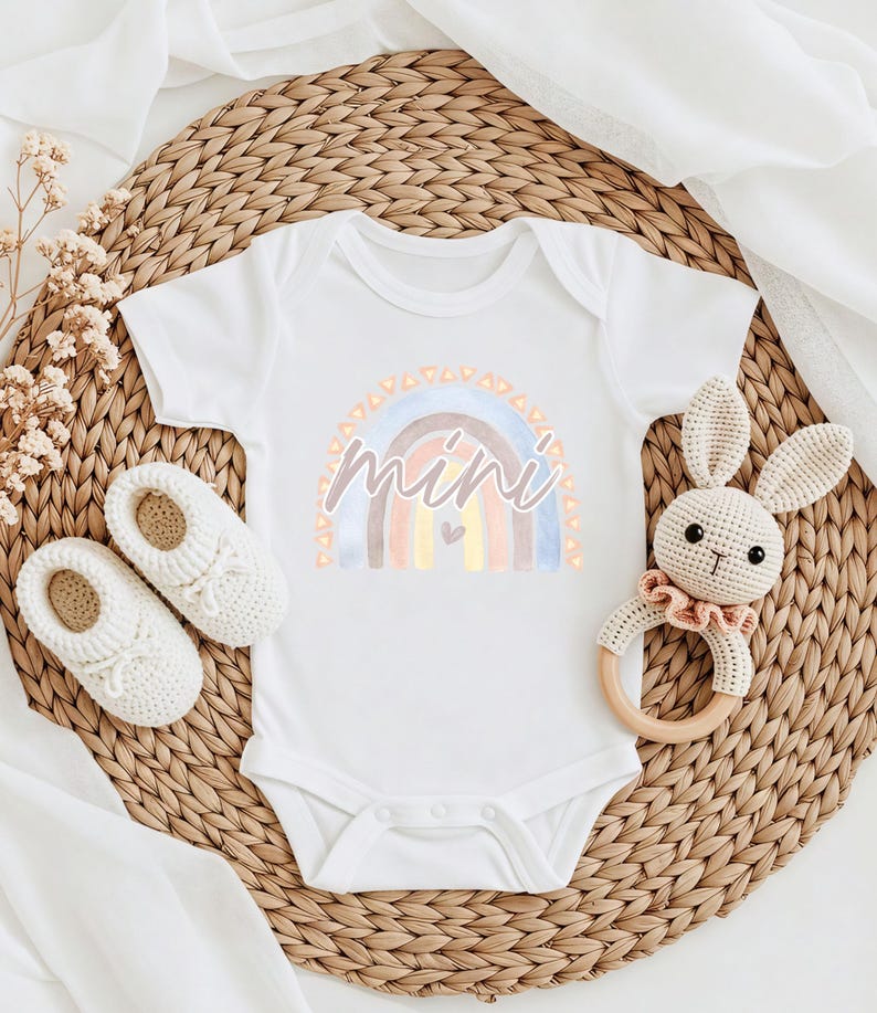 May include: White baby onesie featuring a watercolour rainbow design and the word "mini" in a pink script font. The rainbow has a small heart in the centre.