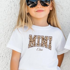 May include: A white t-shirt with a leopard print "MINI" design and the name "Eleni" printed below. A young girl is wearing the shirt and has on black sunglasses.