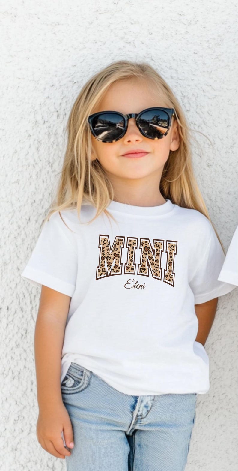 May include: A white t-shirt with a leopard print "MINI" design and the name "Eleni" printed below. A young girl is wearing the shirt and has on black sunglasses.