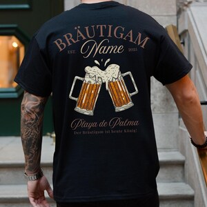 Puede incluir: Camiseta negra con un dise&ntilde;o de tem&aacute;tica cervecera. El dise&ntilde;o presenta dos jarras de cerveza chocando, con las palabras "BR&Auml;UTIGAM Name EST. 2025" y "Playa de Palma" impresas encima y debajo de las jarras. Tambi&eacute;n incluye la frase "Der Br&auml;utigam ist heute K&ouml;nig!"