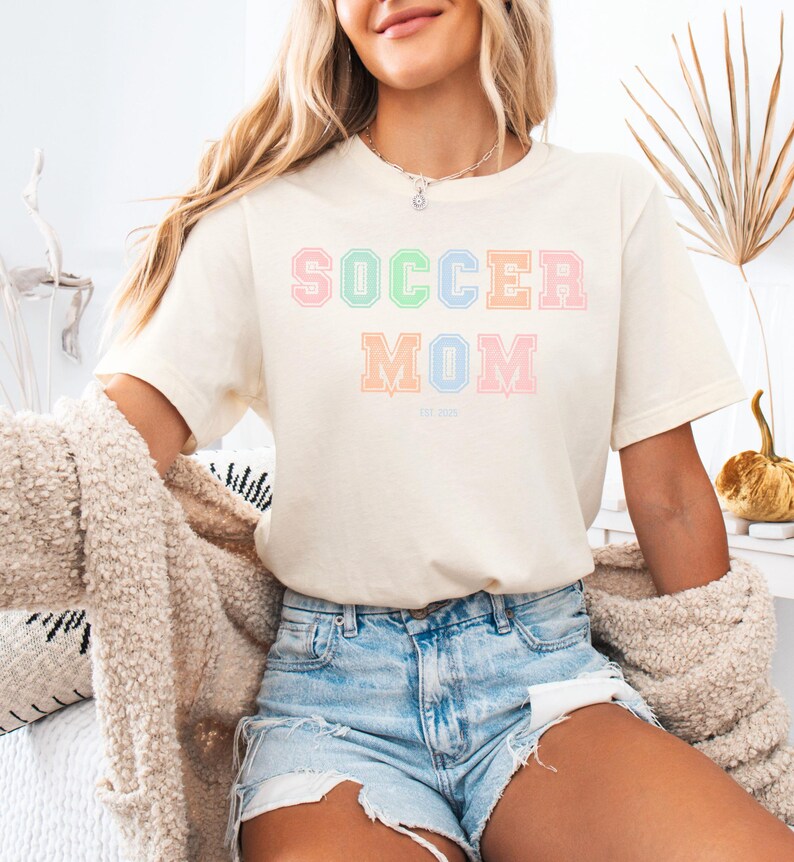 May include: A white t-shirt with the text "SOCCER MOM" in pastel colors. The letters are outlined in a slightly darker shade of the same color. The text "EST. 2022" is printed in a smaller font below the main text.