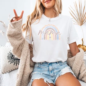 May include: White t-shirt featuring a rainbow design and the word "mama" in a script font. The rainbow is made up of five colours: orange, yellow, green, blue, and purple. The rainbow has a small heart in the centre.