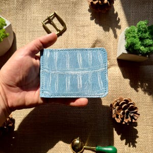 May include: A light blue wallet featuring a textured crocodile skin pattern with visible stitching along the edges. The rectangular wallet has a slim profile and a consistent matte finish across its surface, showcasing a detailed reptilian scale design.