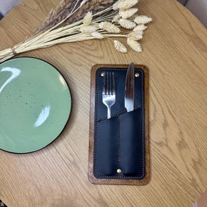 Peut inclure: Un porte-couverts en bois et cuir noir avec une fourchette et un couteau. Le support est rectangulaire avec un cadre en bois marron foncé et des rivets dorés. Une assiette vert clair et des fleurs séchées sont également dans l'image.