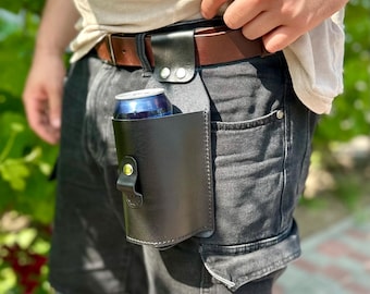 May include: Black leather beer can holder attached to a brown belt. The holder is secured with a snap closure and holds a blue and silver can. The belt is worn with black shorts and a light-colored shirt.