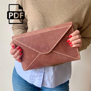 May include: A brown leather envelope clutch with white stitching. The clutch is held by a person wearing a tan sweater, a light blue shirt, and blue jeans. The person has red nail polish. A PDF icon is in the upper left corner.