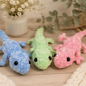 Può includere: Tre lucertole giocattolo di peluche realizzate in morbido filato testurizzato nei colori azzurro, verde pallido e rosa tenue. Ogni lucertola presenta una testa arrotondata, una lunga coda, quattro piccole zampe e due occhi neri lucidi.