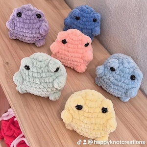 May include: Six small, round, plush frog figures made of soft, chunky yarn in purple, blue, peach, mint green, light blue, and yellow. Each frog features two black plastic eyes. The text @happyknotcreations is visible in the bottom right corner.