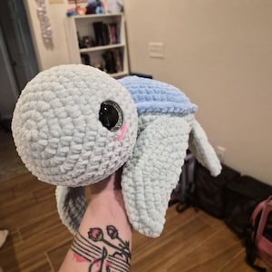 May include: A plush turtle toy crafted from soft, light blue and mint green yarn. The turtle features a large, rounded head with a textured, crocheted surface, oversized black glossy eyes with silver glitter accents, and a small pink stitched mouth.