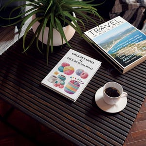 Può includere: Un tavolo di legno marrone scuro con una tazza e un piattino bianchi, una pianta in vaso e due riviste. Una rivista si intitola "TRAVEL MAGAZINE" e l'altra è un "CROCHET GOAL & PROGRESS JOURNAL" con illustrazioni di filato.