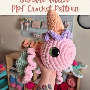 May include: A plush crochet turtle toy with a pink head, a light green shell, and a peach-colored cone on top resembling a sundae. The toy features large black eyes with green glitter accents and ruffled pink and purple yarn trim. Text reads: Sundae Turtle PDF Crochet Pattern.