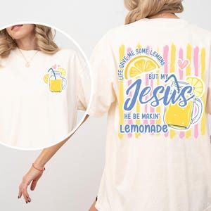 May include: Ivory-colored t-shirt with a lemonade graphic on the front and back. The front features a small lemonade illustration. The back has a larger design with the text "Life gave me some lemons but my Jesus he be makin' lemonade."