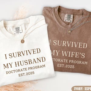 May include: Two ivory and brown t-shirts with the text "I survived my husband's doctorate program est. 2025" and "I survived my wife's doctorate program est. 2025".